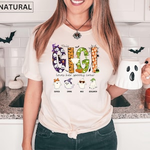 May include: A natural-colored t-shirt featuring a graphic with the text "This GIGI loves her spooky crew" in colorful, patterned letters. Below the text are four cartoon ghosts with names "Sophia", "Emma", "Max", and "Benjamin" printed underneath.