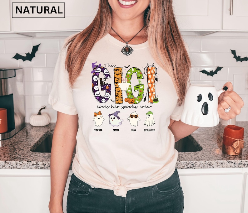 May include: A natural-colored t-shirt featuring a graphic with the text "This GIGI loves her spooky crew" in colorful, patterned letters. Below the text are four cartoon ghosts with names "Sophia", "Emma", "Max", and "Benjamin" printed underneath.
