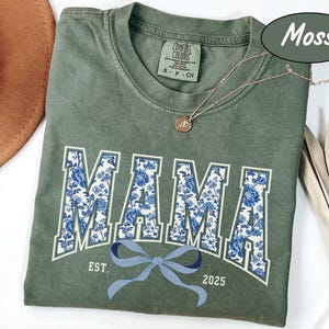 Może przedstawiać: Zielony t-shirt w kolorze mchu z niebieskim i białym nadrukiem kwiatowym, na kt&oacute;rym widnieje napis "MAMA" w stylu uniwersyteckim. Koszulka ma r&oacute;wnież niebieską kokardkę i napis "EST. 2025".