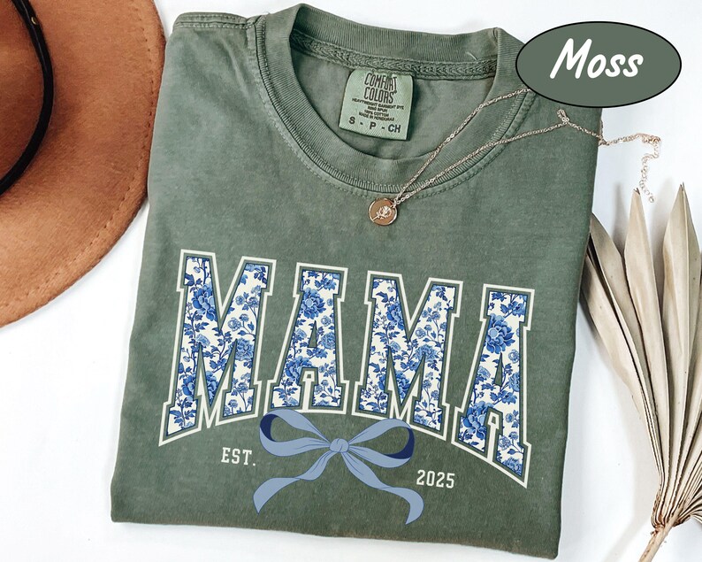 Może przedstawiać: Zielony t-shirt w kolorze mchu z niebieskim i białym nadrukiem kwiatowym, na kt&oacute;rym widnieje napis "MAMA" w stylu uniwersyteckim. Koszulka ma r&oacute;wnież niebieską kokardkę i napis "EST. 2025".