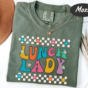 May include: A moss green t-shirt with the words "Lunch Lady" in colorful retro lettering. The letters are outlined in white and have small flowers on them. The shirt has a checkered pattern with small flowers at the top and bottom.