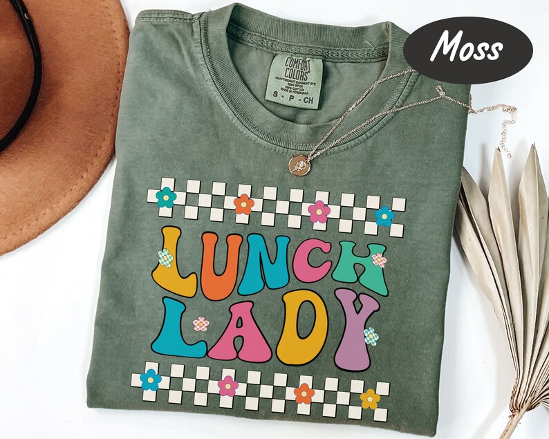May include: A moss green t-shirt with the words "Lunch Lady" in colorful retro lettering. The letters are outlined in white and have small flowers on them. The shirt has a checkered pattern with small flowers at the top and bottom.