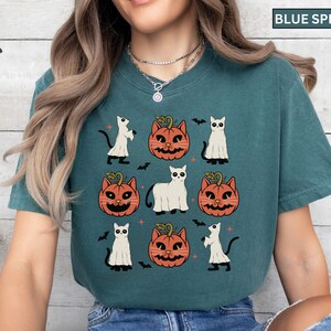 K&ouml;nnte beinhalten: Blaugr&uuml;nes T-Shirt mit einem Halloween-Design. Das Design zeigt orangefarbene K&uuml;rbis-Katzen-Gesichter, wei&szlig;e Geisterfiguren, schwarze Flederm&auml;use und wei&szlig;e Katzenfiguren. Das T-Shirt hat einen l&auml;ssigen Stil.