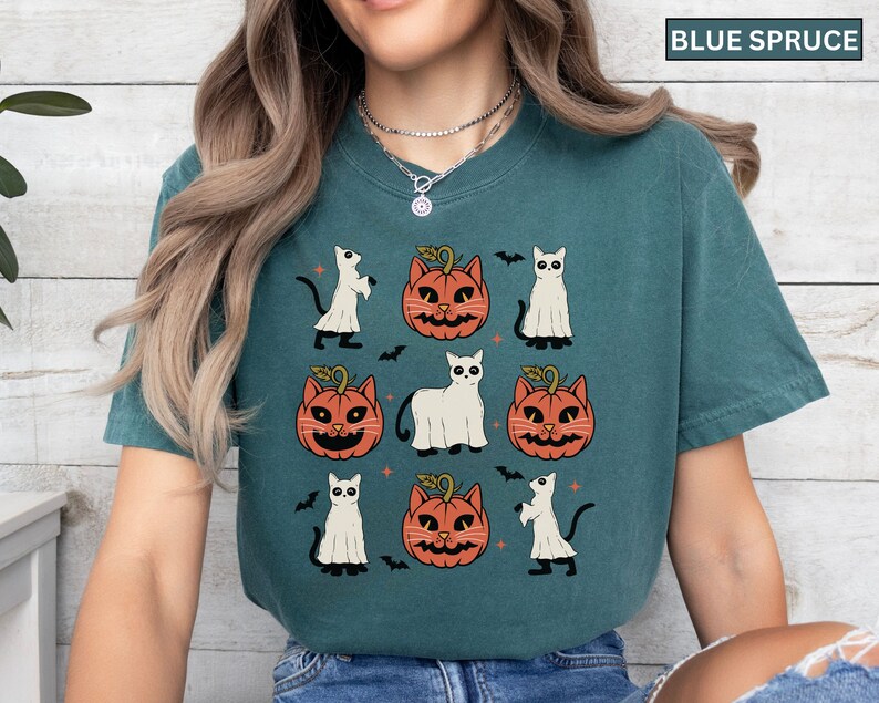 K&ouml;nnte beinhalten: Blaugr&uuml;nes T-Shirt mit einem Halloween-Design. Das Design zeigt orangefarbene K&uuml;rbis-Katzen-Gesichter, wei&szlig;e Geisterfiguren, schwarze Flederm&auml;use und wei&szlig;e Katzenfiguren. Das T-Shirt hat einen l&auml;ssigen Stil.
