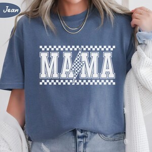 May include: Blue jean colored t-shirt with the word "MAMA" in white, outlined with a checkered pattern. A white lightning bolt graphic is in the center of the word. The text "Blue Jean" is in a blue circle in the upper left corner.