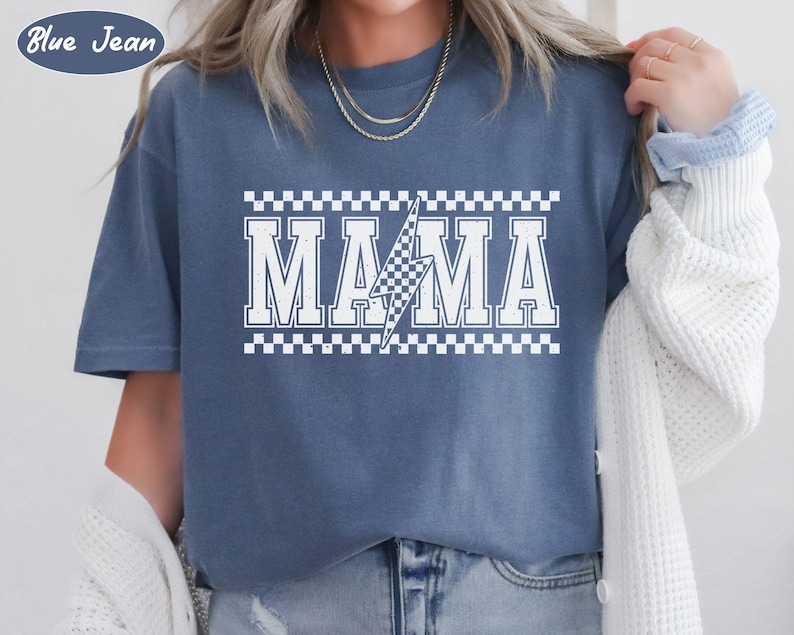 May include: Blue jean colored t-shirt with the word "MAMA" in white, outlined with a checkered pattern. A white lightning bolt graphic is in the center of the word. The text "Blue Jean" is in a blue circle in the upper left corner.