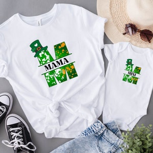 Puede incluir: Dos camisetas blancas con un dise&ntilde;o de tr&eacute;bol verde y sombrero de duende. La camiseta m&aacute;s grande dice "MAMA" y la m&aacute;s peque&ntilde;a dice "MINI".