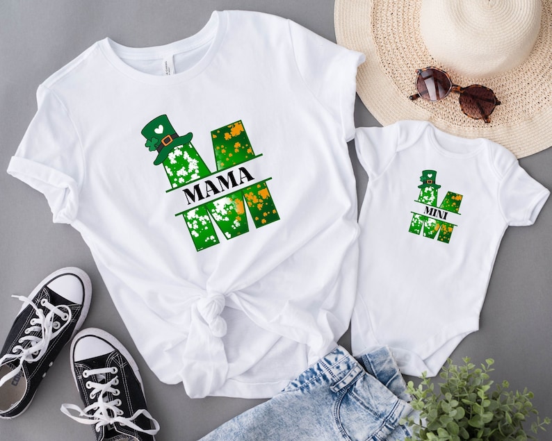 Puede incluir: Dos camisetas blancas con un dise&ntilde;o de tr&eacute;bol verde y sombrero de duende. La camiseta m&aacute;s grande dice "MAMA" y la m&aacute;s peque&ntilde;a dice "MINI".