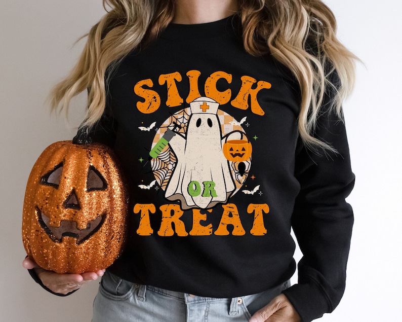 May include: Black sweatshirt with a Halloween graphic. The design shows a ghost in a nurse's cap, holding a syringe and a jack-o'-lantern bucket. The words "STICK OR TREAT" are in orange and green, with bats and a spider web.