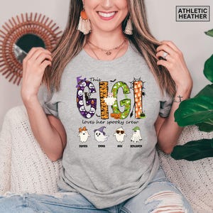 May include: An athletic heather gray t-shirt featuring a graphic with the text "This GIGI loves her spooky crew". The design includes colorful patterned letters, four ghost characters with names underneath, and a small "ATHLETIC HEATHER" label.