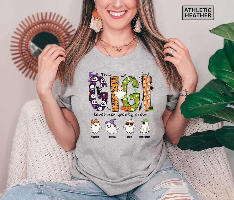 May include: An athletic heather gray t-shirt featuring a graphic with the text "This GIGI loves her spooky crew". The design includes colorful patterned letters, four ghost characters with names underneath, and a small "ATHLETIC HEATHER" label.