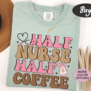 May include: A light blue t-shirt with the text "Half Nurse Half Coffee" in pink and brown retro font. A small pink heart with a stethoscope is above the text. A coffee cup with a pink bow is to the right of the text.