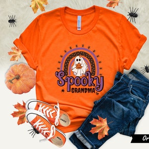 May include: Orange t-shirt with a Halloween-themed design. The graphic features a ghost holding a pumpkin, a rainbow, and the words "Spooky Grandma." The shirt is paired with blue jeans and orange sneakers, with fall leaves and a pumpkin.