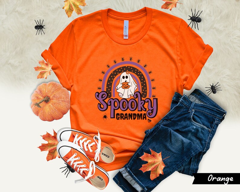 May include: Orange t-shirt with a Halloween-themed design. The graphic features a ghost holding a pumpkin, a rainbow, and the words "Spooky Grandma." The shirt is paired with blue jeans and orange sneakers, with fall leaves and a pumpkin.