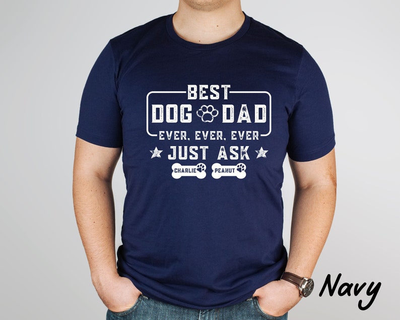 May include: Navy blue t-shirt with white text that reads "BEST DOG DAD EVER. EVER. EVER. JUST ASK" with a paw print graphic. Below the text are two dog bone graphics with the names "CHARLIE" and "PEANUT".