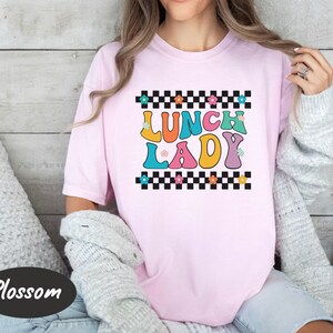 May include: Pink t-shirt with a checkered background and the text "Lunch Lady" in colorful, retro-style letters.