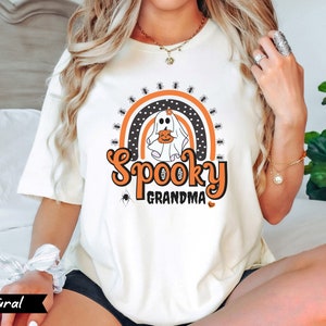May include: Cream-colored t-shirt with a Halloween-themed graphic. The design features a ghost holding a pumpkin, a rainbow, spiders, and the words "Spooky Grandma" in orange and black. The word "Natural" is printed in a black banner at the bottom left.