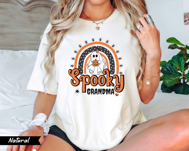 May include: Cream-colored t-shirt with a Halloween-themed graphic. The design features a ghost holding a pumpkin, a rainbow, spiders, and the words "Spooky Grandma" in orange and black. The word "Natural" is printed in a black banner at the bottom left.