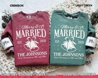 Può includere: Due felpe girocollo color cremisi e verde chiaro, ciascuna con una scritta bianca stampata che recita Merry & Married, Est. 2026, The Johnsons, First Christmas as Mr. and Mrs. e un disegno grafico di due campane.