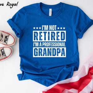 May include: Royal blue t-shirt with white text that reads "I'M NOT RETIRED I'M A PROFESSIONAL GRANDPA". The shirt is displayed with a pair of star-patterned sneakers and a folded American flag.
