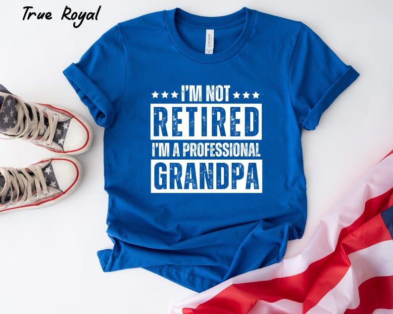 May include: Royal blue t-shirt with white text that reads "I'M NOT RETIRED I'M A PROFESSIONAL GRANDPA". The shirt is displayed with a pair of star-patterned sneakers and a folded American flag.