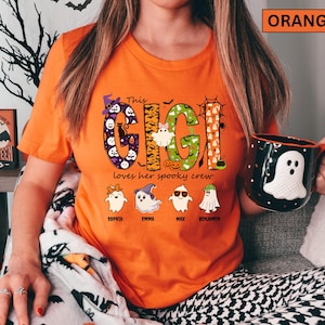 May include: An orange t-shirt featuring a graphic with the text "This GIGI loves her spooky crew". The design includes large letters filled with Halloween patterns, ghosts, pumpkins, and bats, along with four small ghost characters labeled with names.