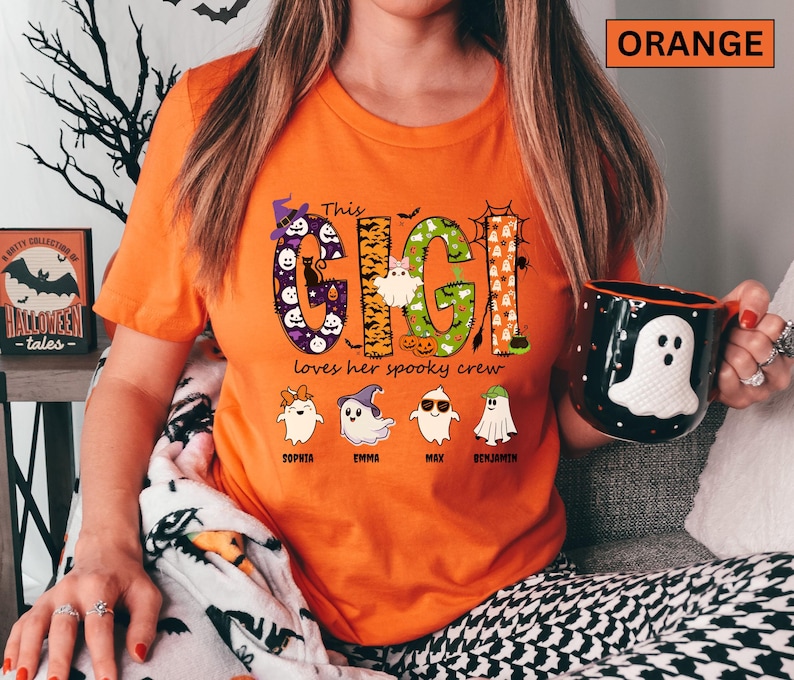 May include: An orange t-shirt featuring a graphic with the text "This GIGI loves her spooky crew". The design includes large letters filled with Halloween patterns, ghosts, pumpkins, and bats, along with four small ghost characters labeled with names.