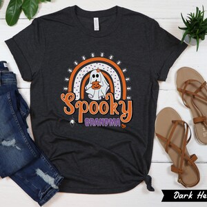 May include: Dark heather gray t-shirt with a Halloween-themed graphic. The design features a ghost holding a pumpkin, a rainbow, and the words "Spooky Grandma" in orange and purple. The shirt is styled with blue jeans and brown sandals.