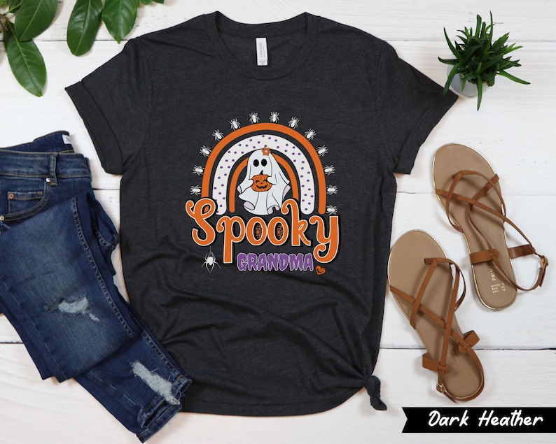 May include: Dark heather gray t-shirt with a Halloween-themed graphic. The design features a ghost holding a pumpkin, a rainbow, and the words "Spooky Grandma" in orange and purple. The shirt is styled with blue jeans and brown sandals.