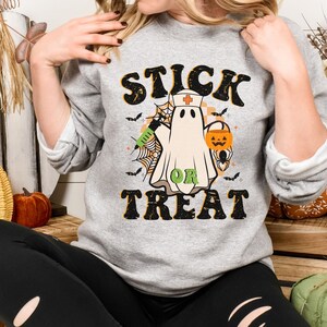 May include: A grey sweatshirt with a Halloween graphic. The design features a ghost in a nurse's cap, holding a syringe and a jack-o'-lantern, with the words "STICK OR TREAT" in a retro font. Bats and spiderwebs complete the design.