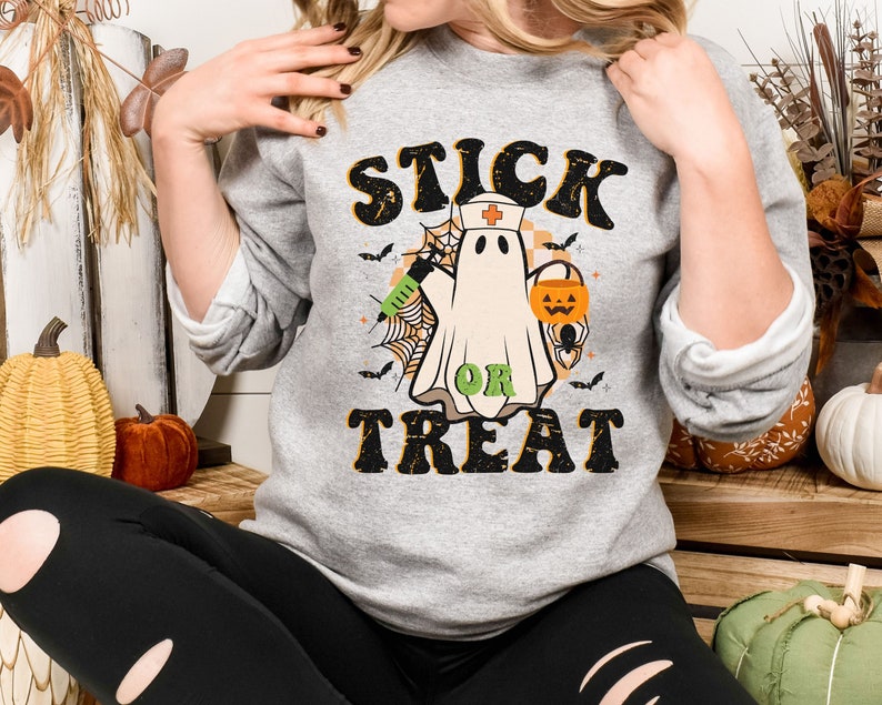 May include: A grey sweatshirt with a Halloween graphic. The design features a ghost in a nurse's cap, holding a syringe and a jack-o'-lantern, with the words "STICK OR TREAT" in a retro font. Bats and spiderwebs complete the design.