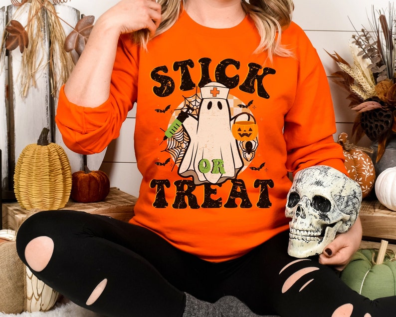May include: Bright orange long-sleeved sweatshirt with a Halloween design. The graphic shows a ghost in a nurse's hat, holding a syringe and a pumpkin lantern, with the words "STICK OR TREAT". Black bats and a spider's web are also included.