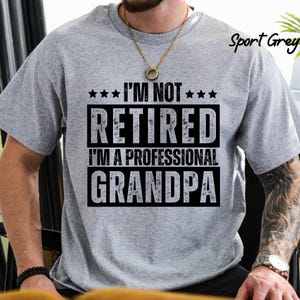 May include: Sport Grey t-shirt with the text "I'M NOT RETIRED I'M A PROFESSIONAL GRANDPA" in black. The shirt has a crew neck and short sleeves. The text is in a bold, blocky font.