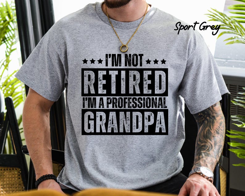 May include: Sport Grey t-shirt with the text "I'M NOT RETIRED I'M A PROFESSIONAL GRANDPA" in black. The shirt has a crew neck and short sleeves. The text is in a bold, blocky font.