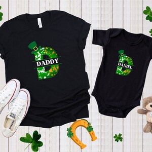 Puede incluir: Camiseta y body negros con un dise&ntilde;o de tr&eacute;bol verde y sombrero de duende. La camiseta dice "Daddy" y el body dice "Daniel".