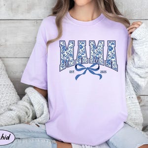 Może przedstawiać: Fioletowy t-shirt z niebieskim i białym nadrukiem kwiatowym, na kt&oacute;rym widnieje napis "MAMA EST. 2025" z niebieską kokardką.