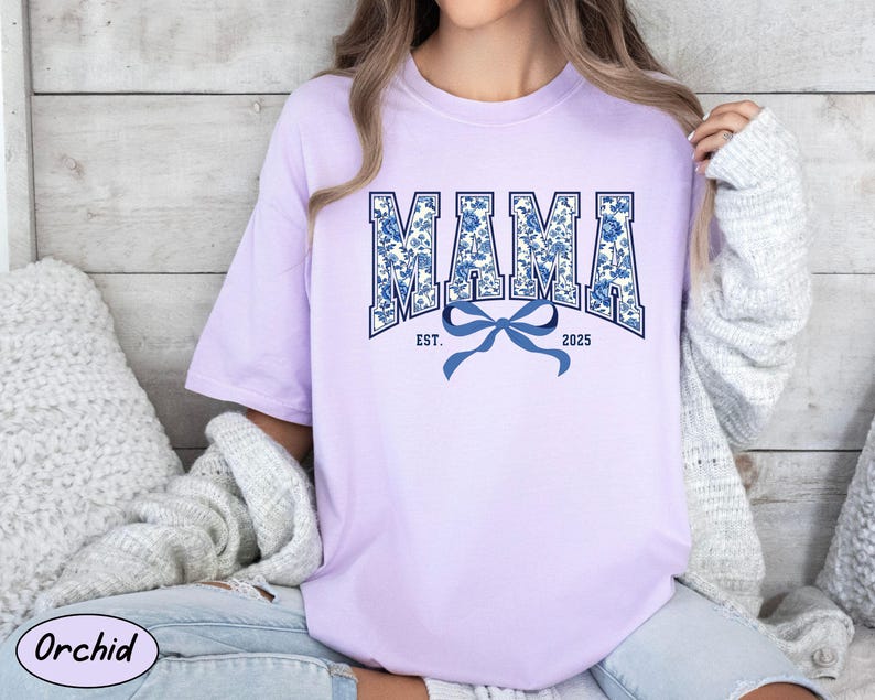 Może przedstawiać: Fioletowy t-shirt z niebieskim i białym nadrukiem kwiatowym, na kt&oacute;rym widnieje napis "MAMA EST. 2025" z niebieską kokardką.
