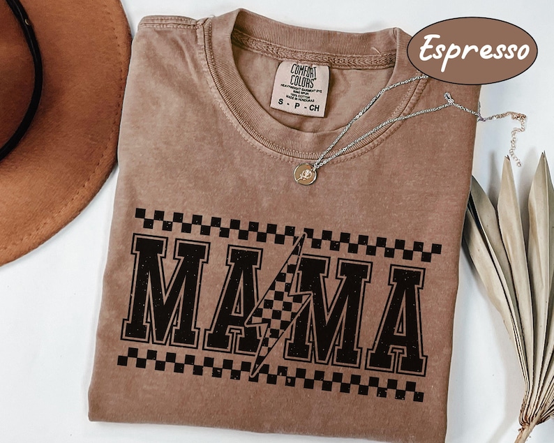 May include: Espresso-colored t-shirt with the word "MAMA" in a black, distressed, collegiate-style font, with a checkered pattern above and a lightning bolt graphic. The shirt is a Comfort Colors brand, and a silver necklace with a pendant is visible.