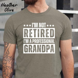 May include: Heather olive green t-shirt with the text "I'M NOT RETIRED I'M A PROFESSIONAL GRANDPA" in a white, bold font. The shirt has a crew neck and short sleeves. The words "Heather Olive" are printed in the upper left corner.