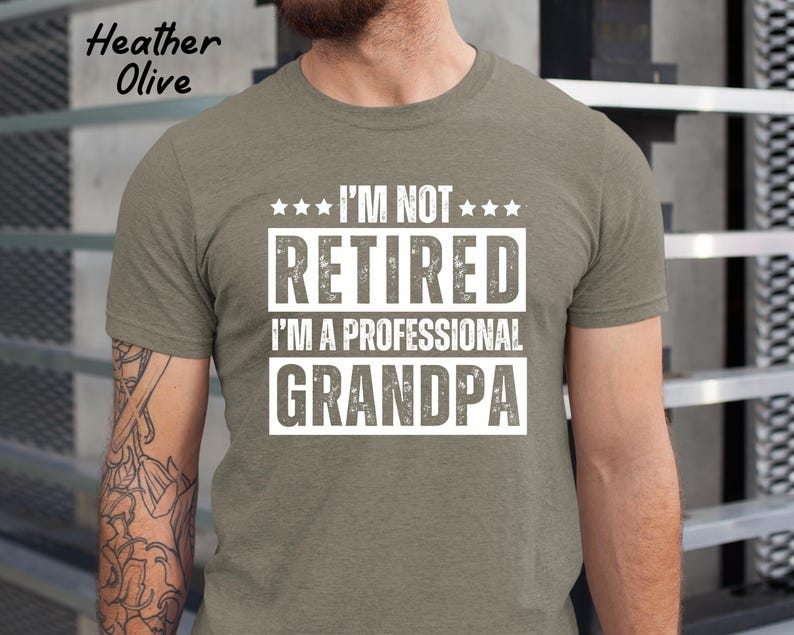 May include: Heather olive green t-shirt with the text "I'M NOT RETIRED I'M A PROFESSIONAL GRANDPA" in a white, bold font. The shirt has a crew neck and short sleeves. The words "Heather Olive" are printed in the upper left corner.