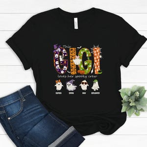 May include: A black short-sleeved t-shirt featuring the text "This GIGI loves her spooky crew" in large, patterned letters. Below the text are four cartoon ghosts with names "Sophia", "Emma", "Max", and "Benjamin" printed underneath each.
