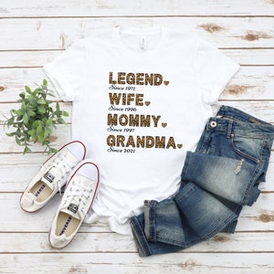 May include: White t-shirt with a leopard print design that says "LEGEND. Since 1971 WIFE. Since 1996 MOMMY. Since 1997 GRANDMA. Since 2021"