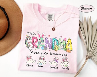 Könnte beinhalten: Hellrosa T-Shirt mit der Aufschrift „This Grandma loves her bunnies“ in bunten Buchstaben und Grafiken. Das Shirt zeigt Hasenillustrationen mit Namen darunter. Eine goldene Halskette ist auf dem Shirt drapiert.