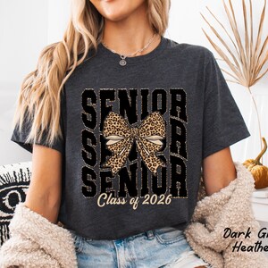 Puede incluir: Camiseta de color gris oscuro jaspeado con un gr&aacute;fico negro de la palabra "Senior" repetida tres veces con un lazo de estampado de leopardo en el centro y el texto "Class of 2026" debajo.