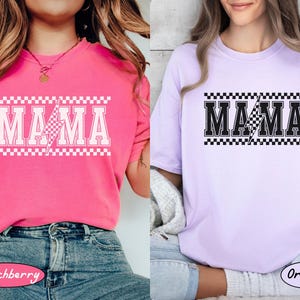 May include: Two short-sleeved t-shirts, one pink and one lavender, each with the word "MAMA" in bold, white, block letters. A lightning bolt graphic is in the center. The word is framed by a checkered pattern.