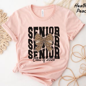 Puede incluir: Una camiseta rosa con un gr&aacute;fico negro que dice "Senior" con un lazo de estampado de leopardo en el medio y "Class of 2026" en la parte inferior.