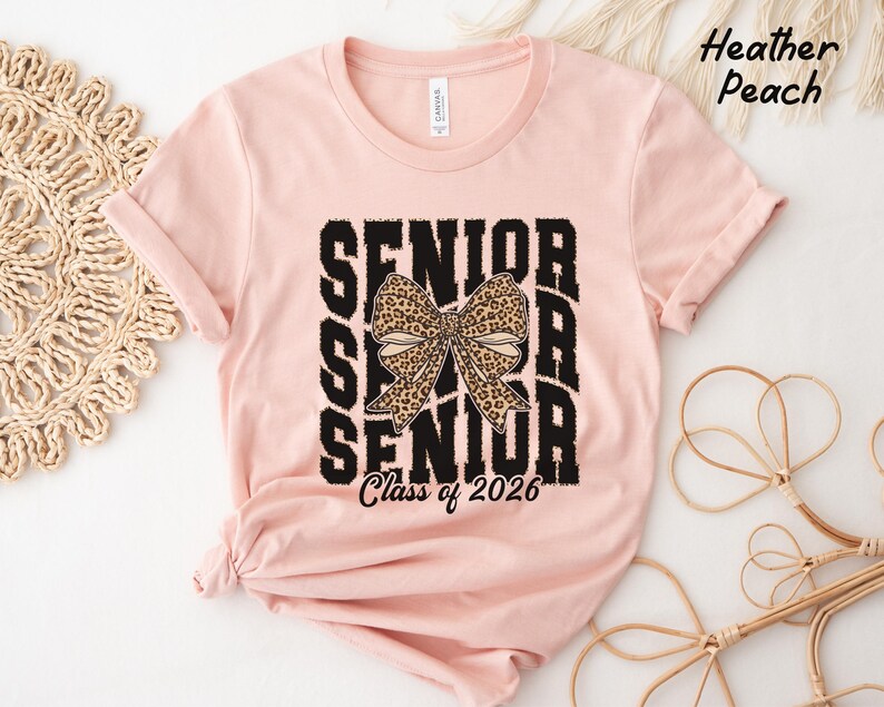 Puede incluir: Una camiseta rosa con un gr&aacute;fico negro que dice "Senior" con un lazo de estampado de leopardo en el medio y "Class of 2026" en la parte inferior.