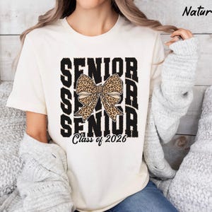 Puede incluir: Una camiseta blanca con un gr&aacute;fico negro que dice "Senior" con un lazo de estampado de leopardo en el medio y "Class of 2026" en la parte inferior.