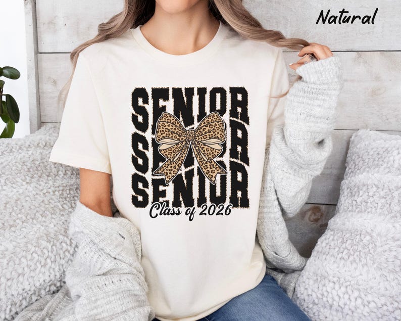 Puede incluir: Una camiseta blanca con un gr&aacute;fico negro que dice "Senior" con un lazo de estampado de leopardo en el medio y "Class of 2026" en la parte inferior.
