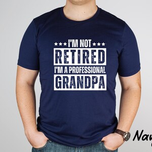 May include: Navy blue t-shirt with the text "I'M NOT RETIRED I'M A PROFESSIONAL GRANDPA" in white. The shirt has a crew neck and short sleeves. The word "Navy" is printed in the bottom right corner.
